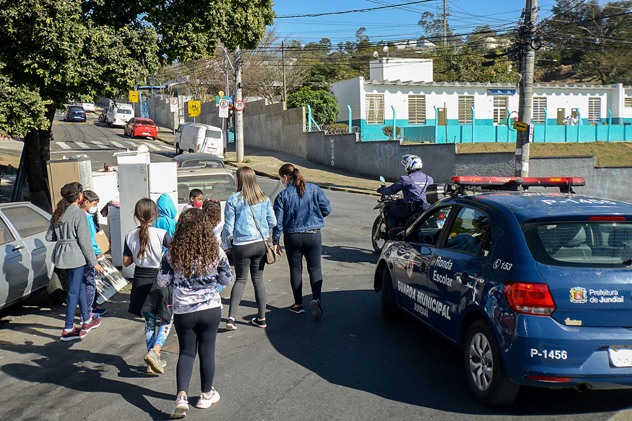 Crianças caminham por uma rua de bairro, junto deles estão uma moto e uma viatura da Guarda Municipal. A viatura é azul, com escritos em branco. Na lateral pode-se ler Ronda Escolar - Guarda Municipal Jundiaí.