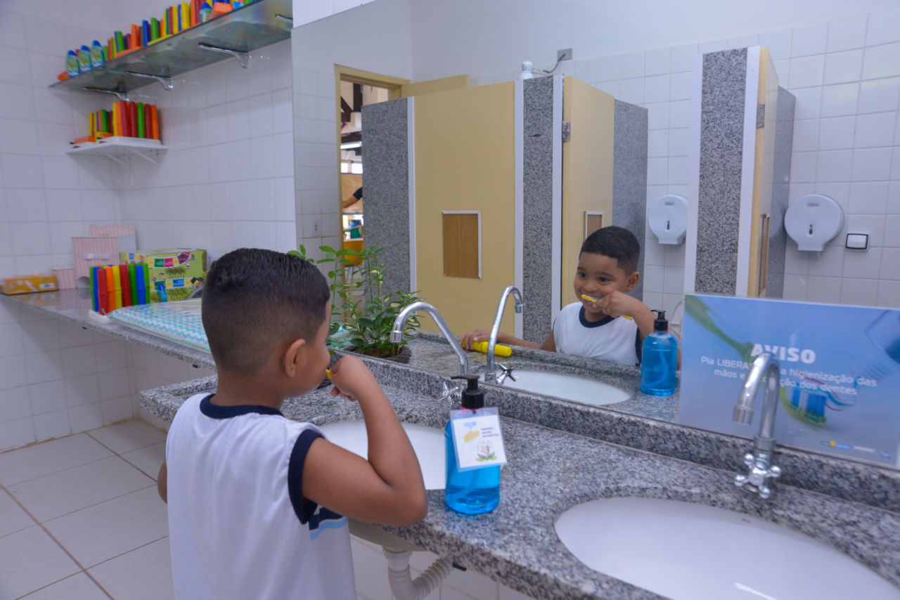 DESCRIÇÃO DA IMAGEM: Garoto com uniforme escolar, está em frente a uma pia, fazendo a escovação de dentes