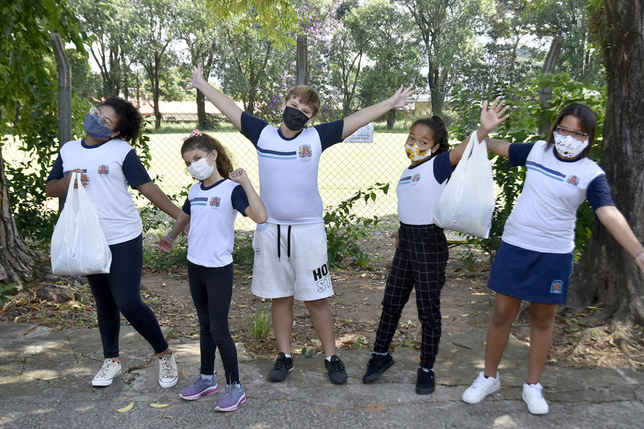 Imagem mostra cinco crianças, que estão com a camiseta do uniforme escolar branca com gola, mangas e detalhes azuis, calça preta, ou bermuda azul e branca, estão com sacolas nas mãos, comemorando