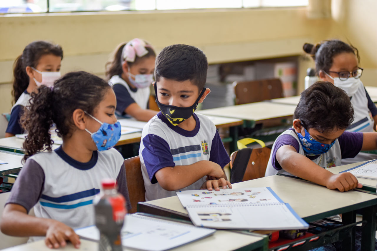 DESCRIÇÃO DA IMAGEM:
Crianças estão dentro de sala de aula, usando uniforme branco com detalhes em azul, usando máscara de proteção facial e folheando livros didáticos.