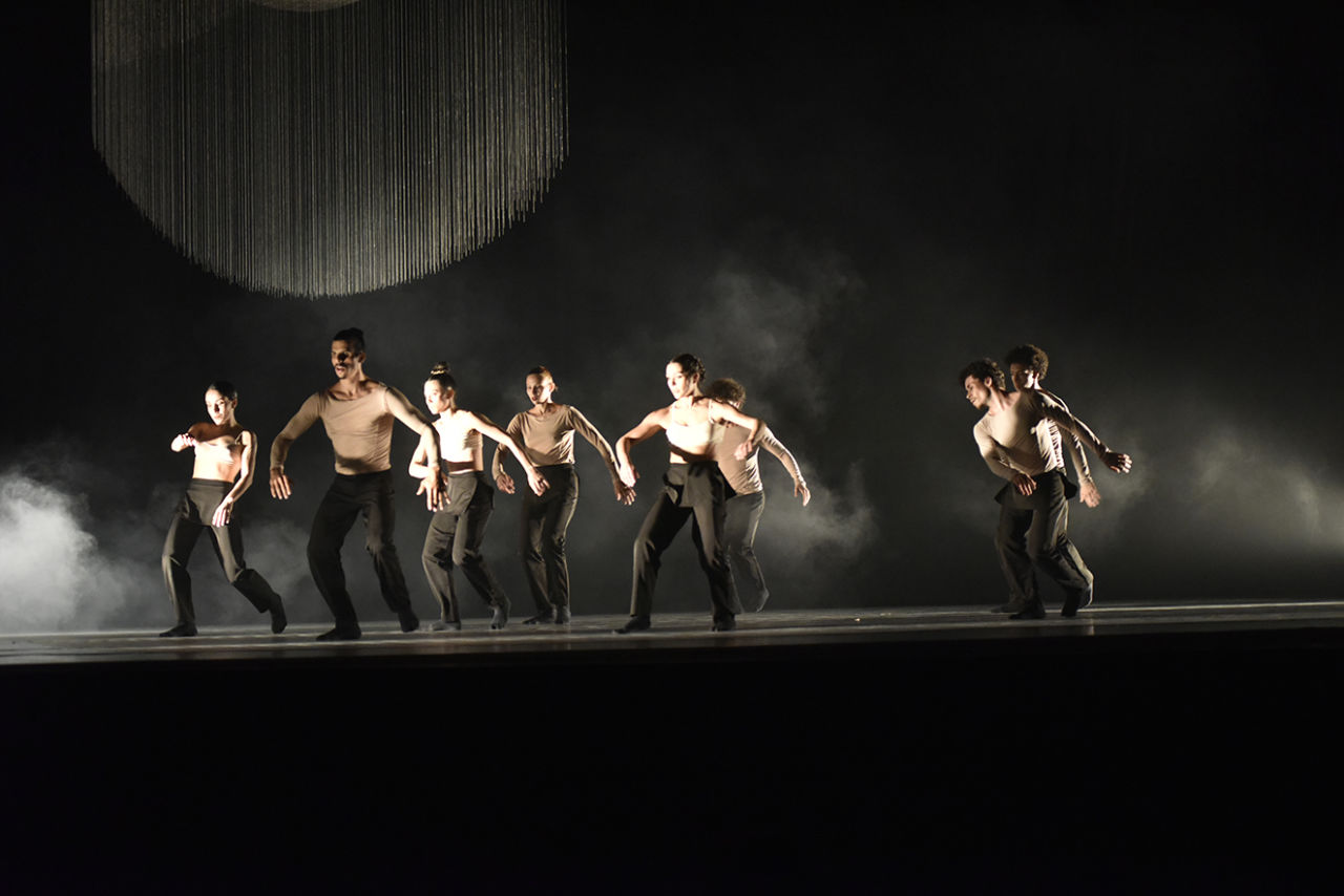 Bailarinos homens e mulheres com calças pretas e parte superior cor bege, dançando sobre palco iluminado com fumaça e enfeites metálicos ao fundo