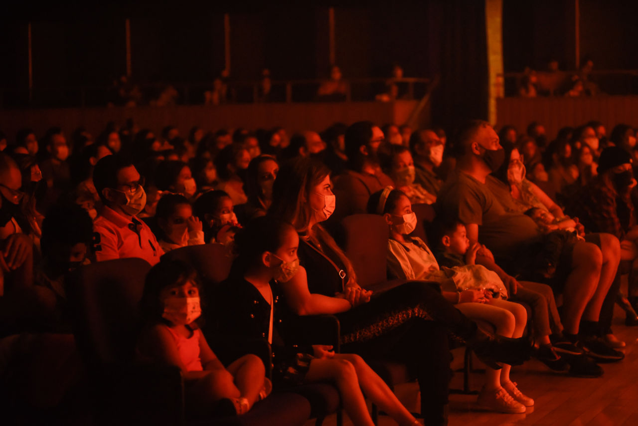 Crianças, mulheres e homens sentados em plateia de teatro com as luzes apagadas e nuance de luzes vermelhas
