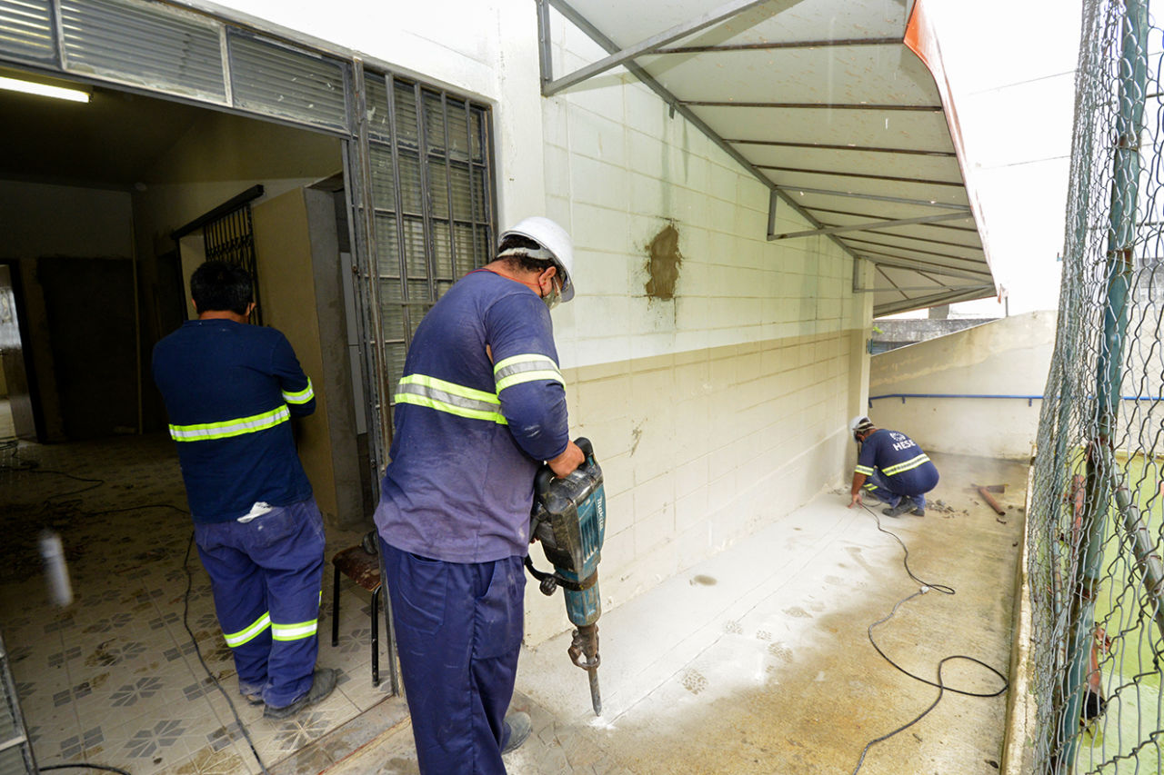 Três homens, de uniforme azul, com fita refletiva, usam equipamentos de segurança e estão trabalhando em obra de prédio. Um deles segura um equipamento azul nas mãos para fazer o corte de cimento.