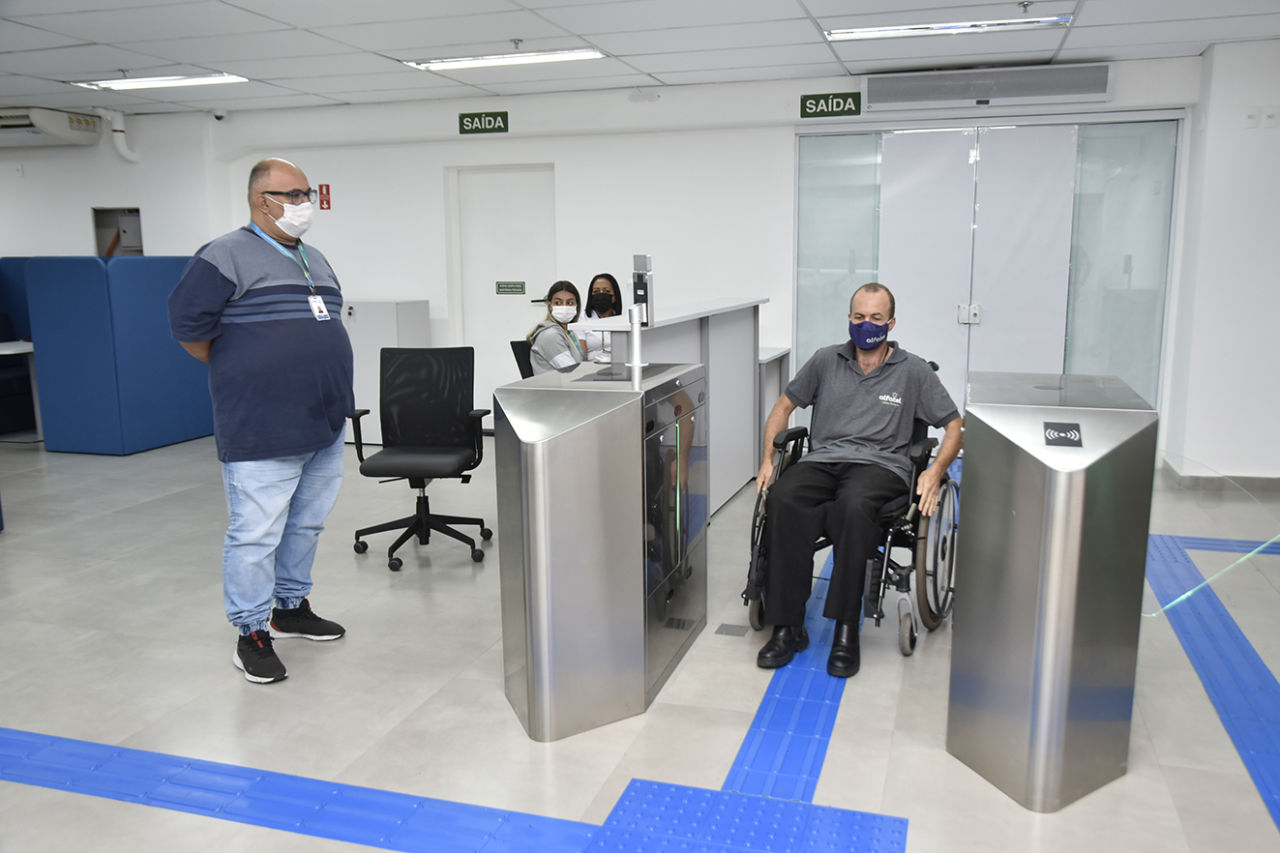 Pessoa com cadeira de rodas com camisa cinza e calça preta entra na recepção do local de máscara observado por servidor em pé com camisa cinza e azul