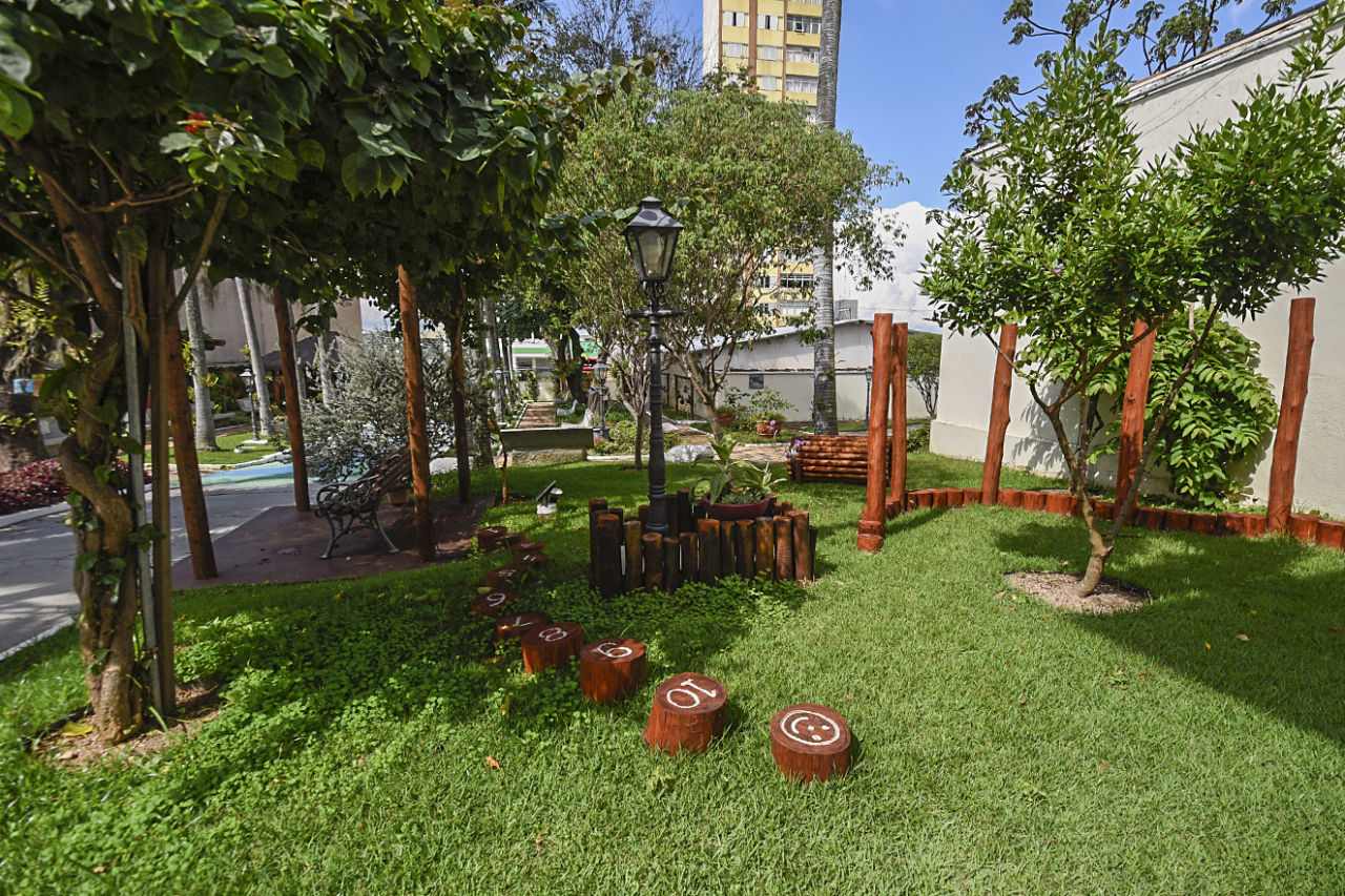 Jardim com gramado, árvores, parede e luminária, com instalação com objetos de madeira no chão