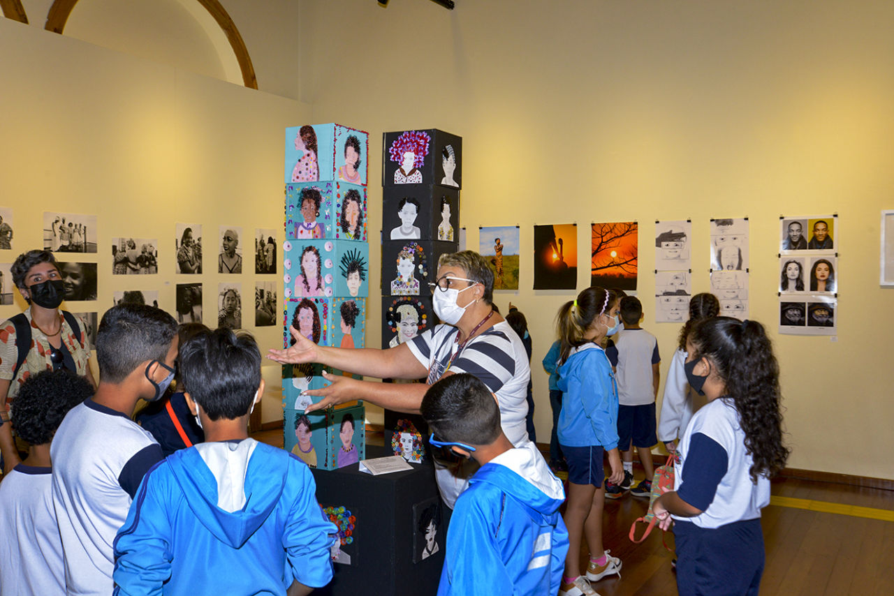 Crianças com uniformes escolares são acompanhadas por adultas em sala de exposição com fotos nas paredes e desenhos de figuras em dois totens, um azul e outro preto