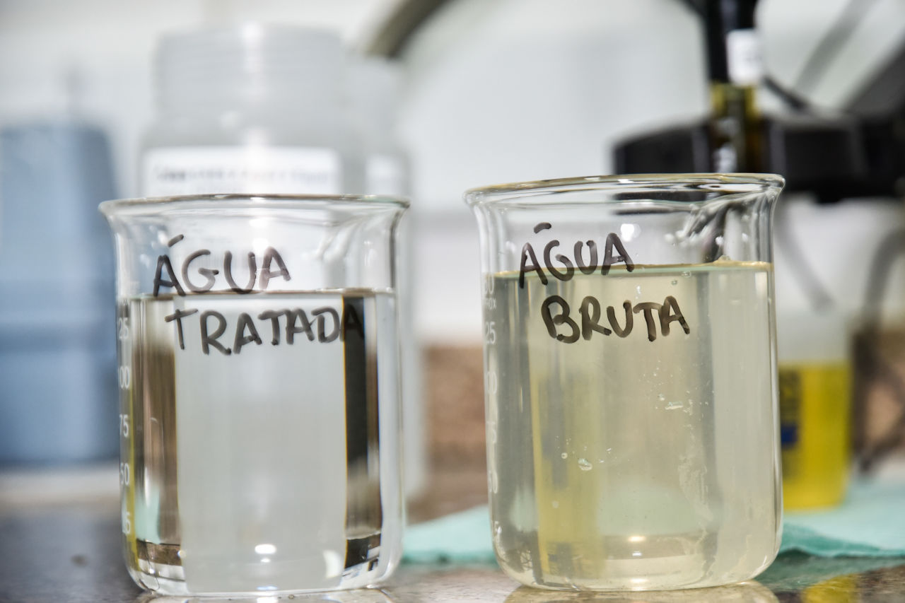 Imagem mostra dois recipientes, um com água tratada e outro com água bruta, no Laboratório