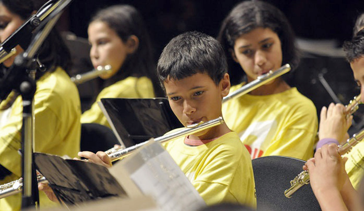 Meninos e meninas com camisetas amarelas, sentadas próximas e tocando instrumentos de sopro com partituras à frente