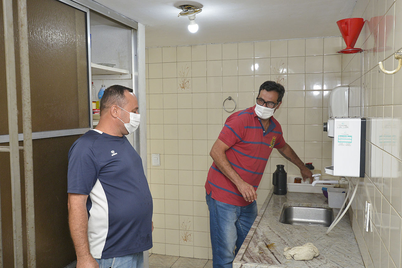 Dois homens usando máscaras em cozinha residencial, um deles usando óculos e mexendo com a louça perto de pia