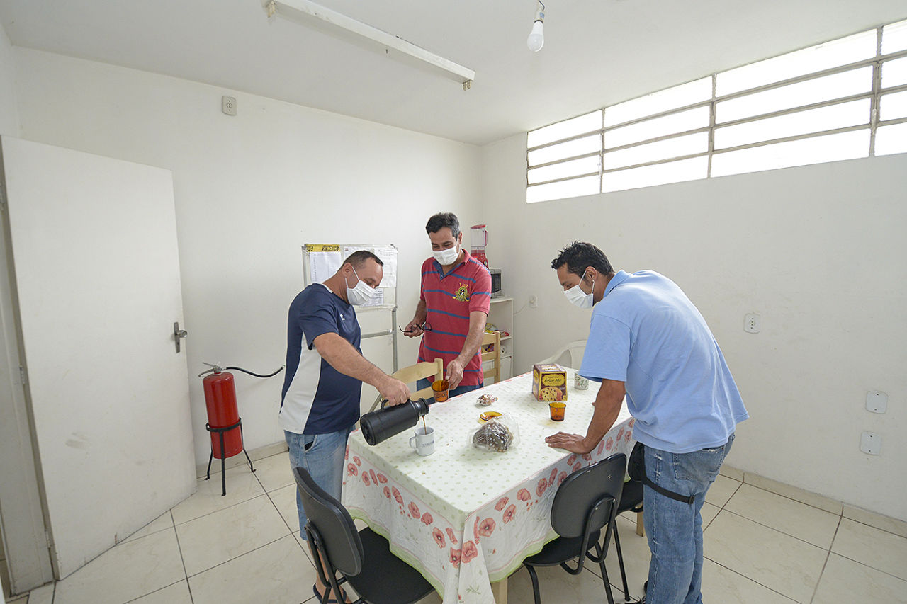 Três homens em pé ao redor de uma mesa retangular, um deles apoiado sobre a mesa, outro segurando uma xícara de vidro e outro colocando café em xícara branca apoiada sobre a mesa, onde também se vê um panetone e outros pequenos pacotes 
