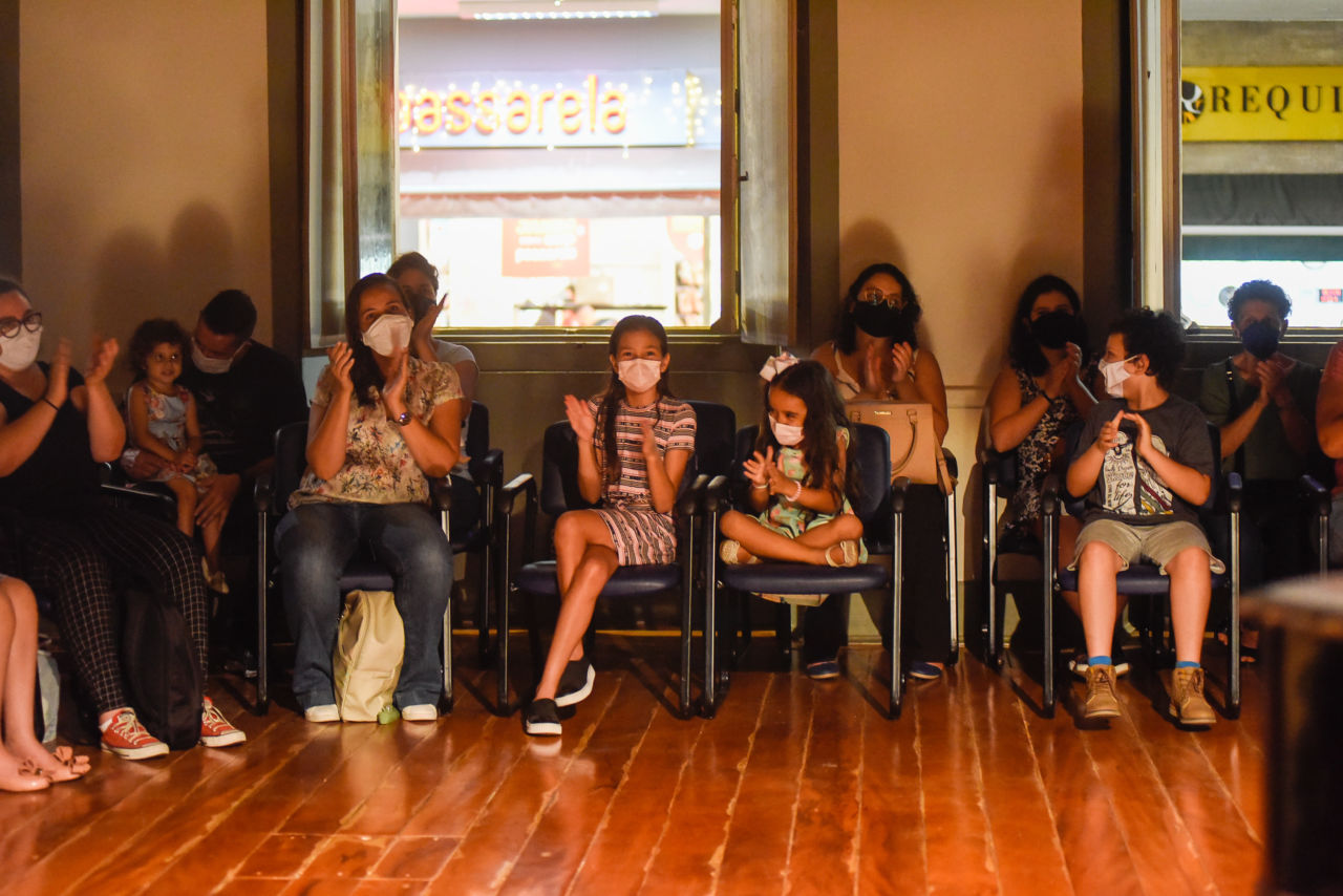 Crianças, mulheres e homens com máscara facial, sentados em cadeiras de sala de espetáculo com piso de madeira e janelas abertas, com letreiros luminosos de comércio ao fundo