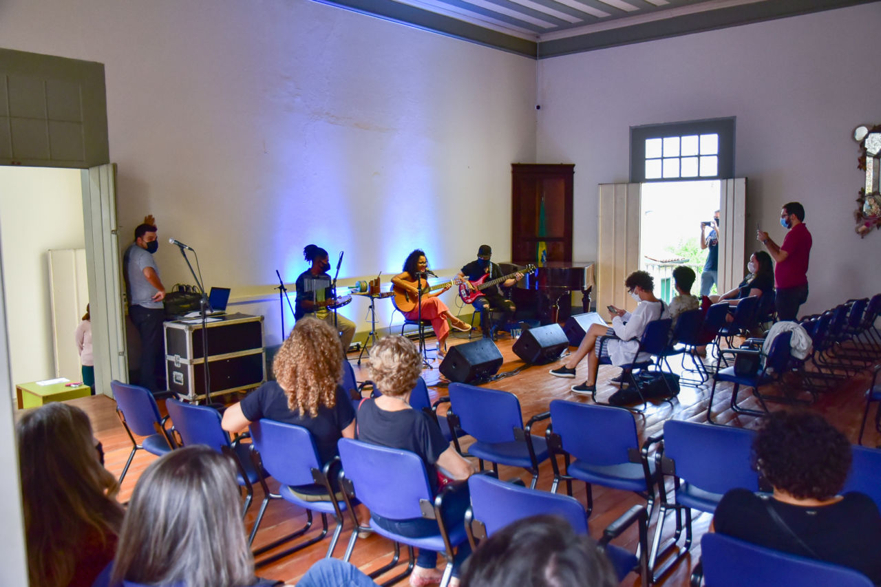 Sala de apresentação com piso e teto de madeira, com trio de músicos tocando percussão, violão e baixo, e plateia sentada em cadeiras de estofado azul