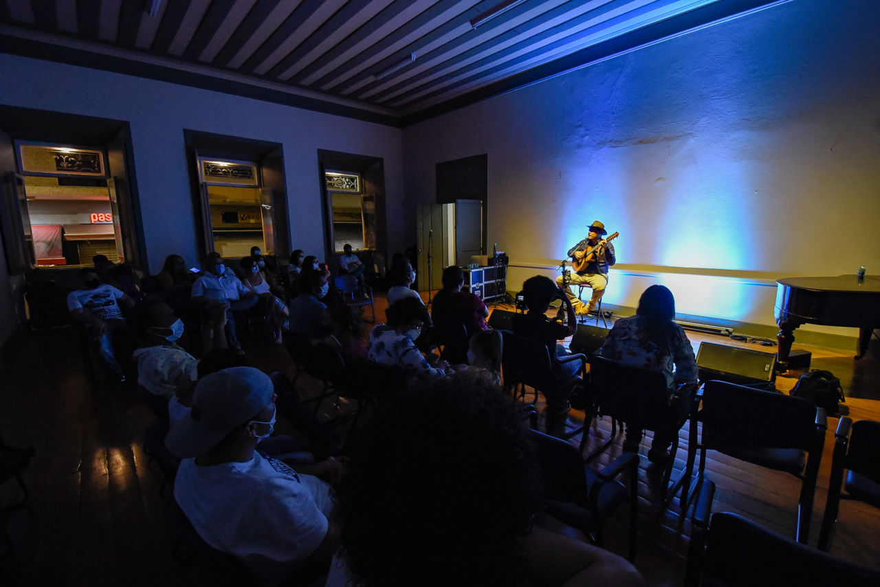 Homem de chapéu tocando violão e cantando no microfone, iluminado, em sala de espetáculo sem iluminação com focos de luz azul 