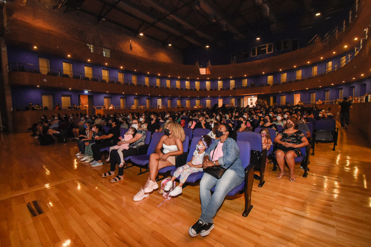 Foto da plateia do Teatro Polytheama com luzes baixas, com crianças, mulheres e homens, todos de máscaras, sentados nas poltronas de forro azul.