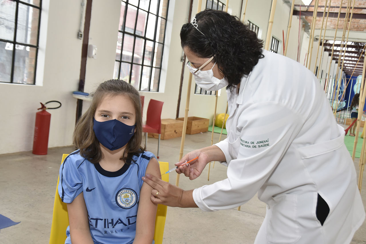Mulher com jaleco branco aplica vacina no braço de uma garota que está com máscara e camiseta azul.