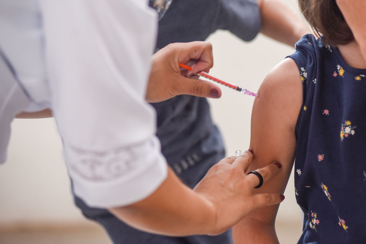 Imagem mostra mão de mulher, aplicando vacina no braço de uma criança.