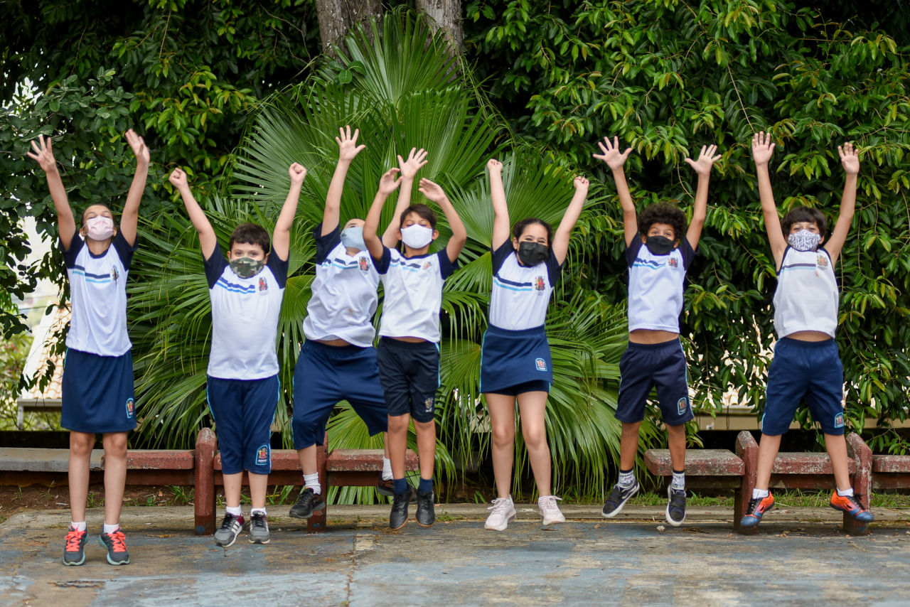 Na imagem há sete crianças pulando com uniforme da Prefeitura de Jundiaí com árvores ao fundo da escola.