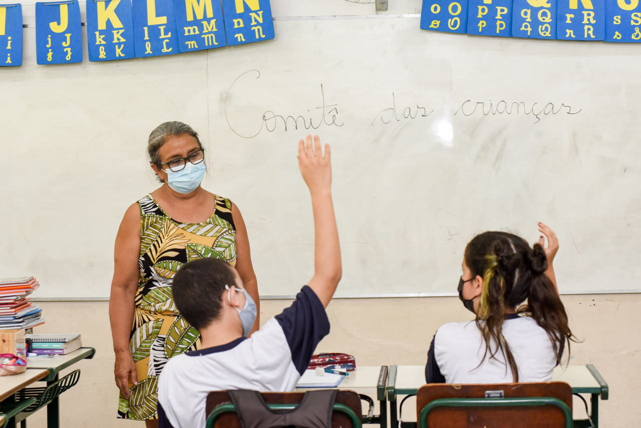 Em sala de aula, ao fundo há uma lousa com o escrito "Comitê das Crianças", na frente a professora olhando para dois alunos, uma menina e um menino sentados com as mãos levantadas.