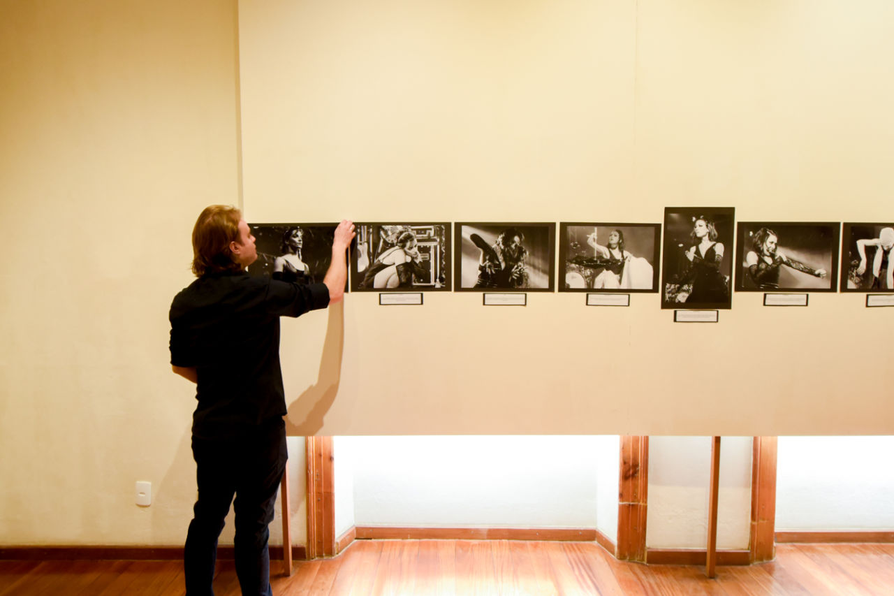 Homem de cabelos loiros lisos, compridos até a altura dos ombros, usando camisa social preta, ajeita fotografias em preto e branco em parede de sala com pavimento de madeira