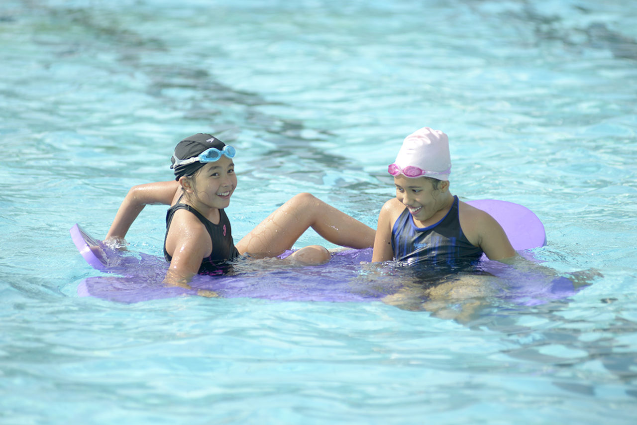 Duas meninas de touca e maiô brincam na piscina