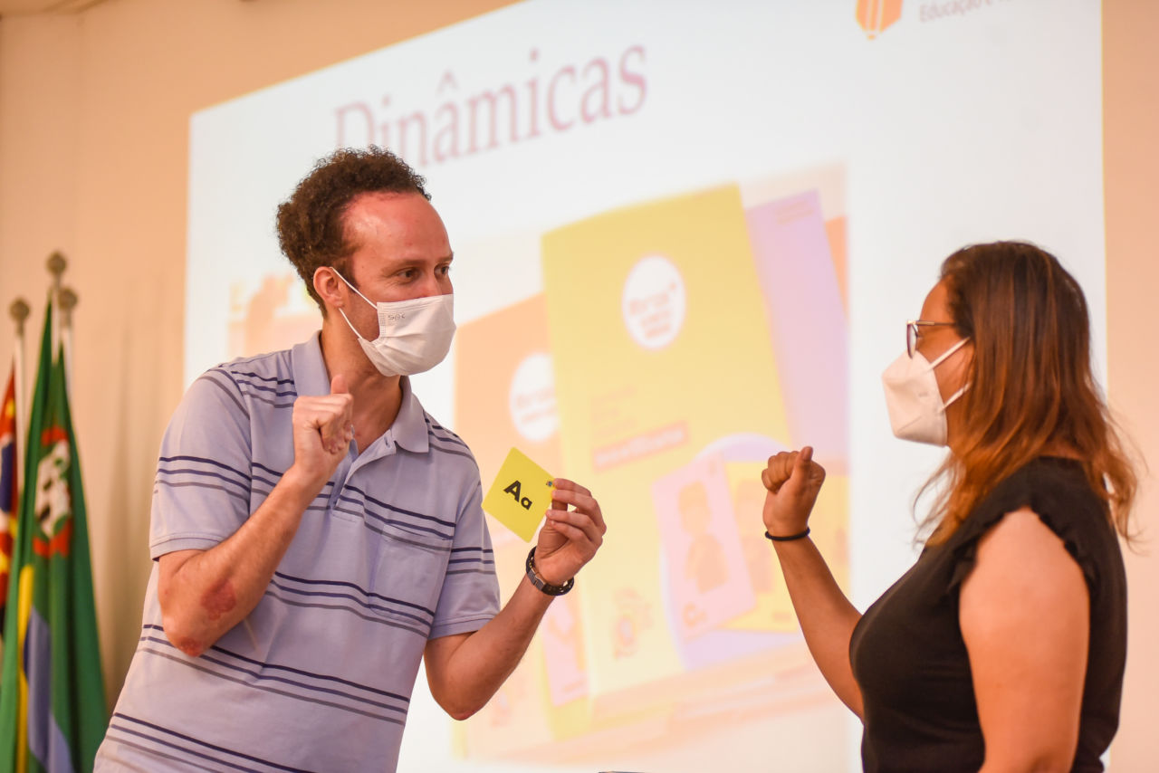 DESCRIÇÃO DA IMAGEM
homem com camisa azul e listras está usando máscara facial e gesticulando, com um cartão amarelo em uma das mãos no qual se lê Aa. Em frente está uma mulher com camisa preta, ela também usa máscara facial e gesticula. 