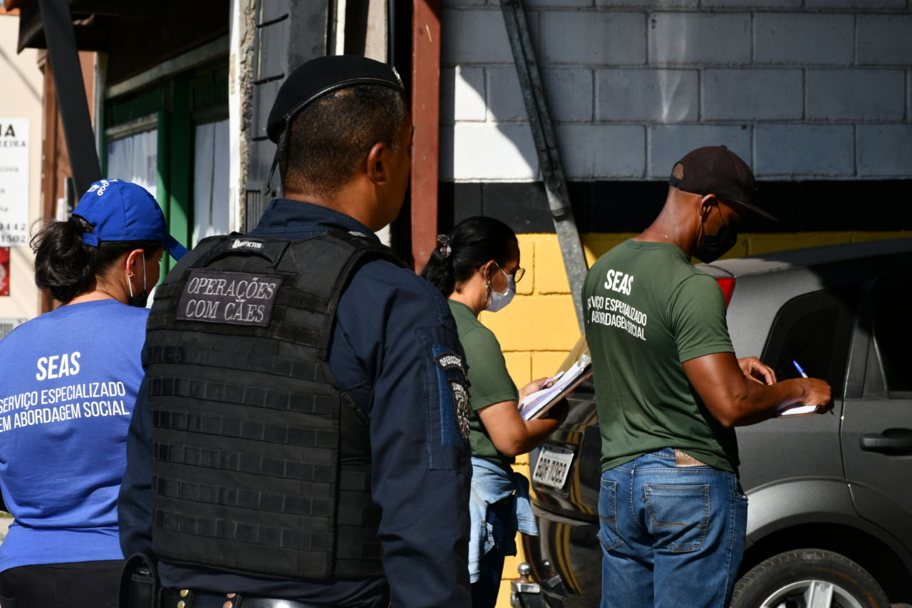 Pessoas com uniforme e guarda municipal em frente a uma garagem de mecânico, segurando prancheta e caneta e conversando com alguém