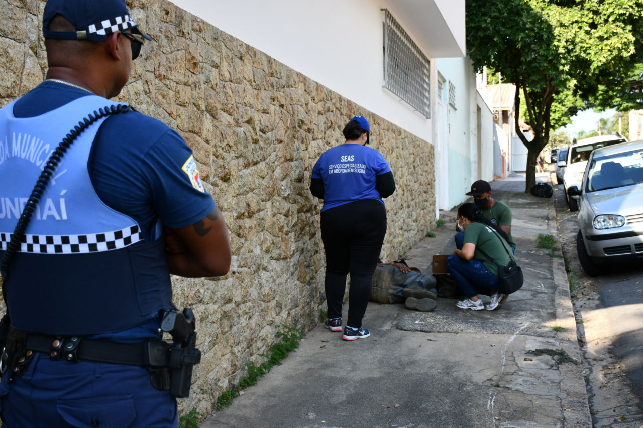 Pessoas com uniforme e guarda municipal abordam pessoa deitada sobre a calçada em rua de residências com carros estacionados e árvores 
