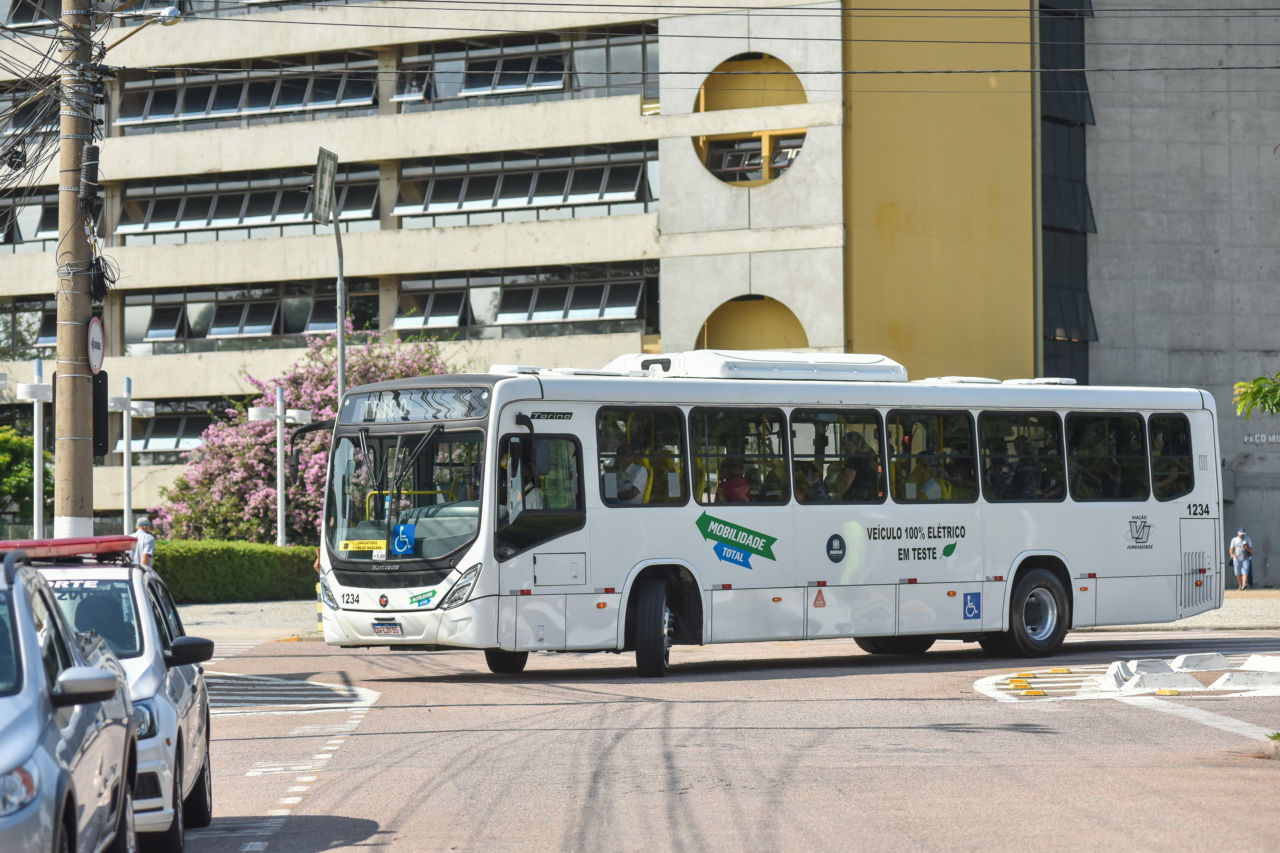 Descrição da Imagem: ônibus pintado de branco, com dizeres 'Mobilidade Total', o brasão da prefeitura de Jundiaí e a indicação de ser um veículo 100% elétrico circula pela avenida da Liberdade, em frente ao Paço Municipal