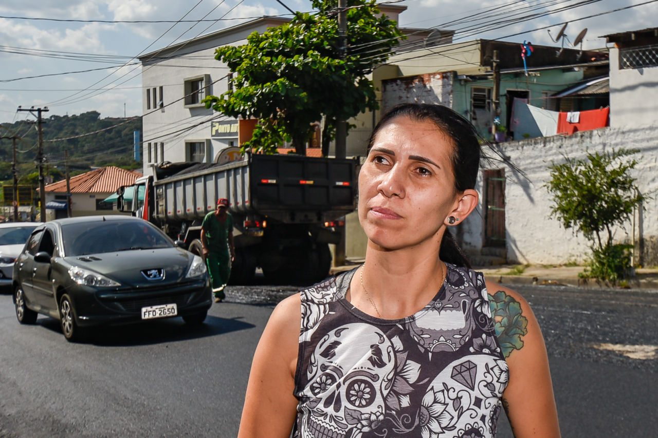 Com uma blusinha preta e branca, Ana Paula tem os cabelos presos, é uma mulher de pele morena e está à frente da rua, com um caminhão e um carro ao fundo