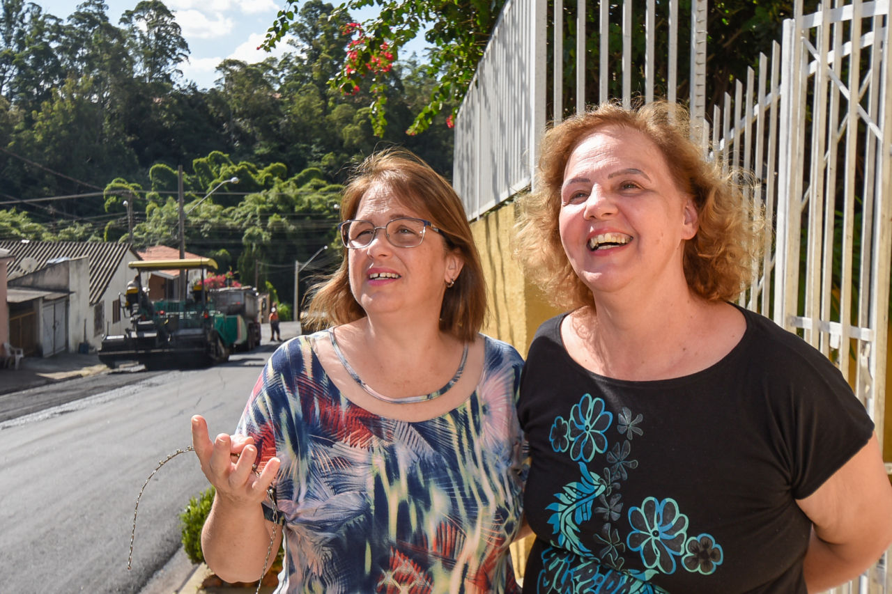 As irmãs Arlete e Ana Lúcia estão sorridentes, em frente ao portão de sua casa e ao lado da rua que recebe o asfalto
