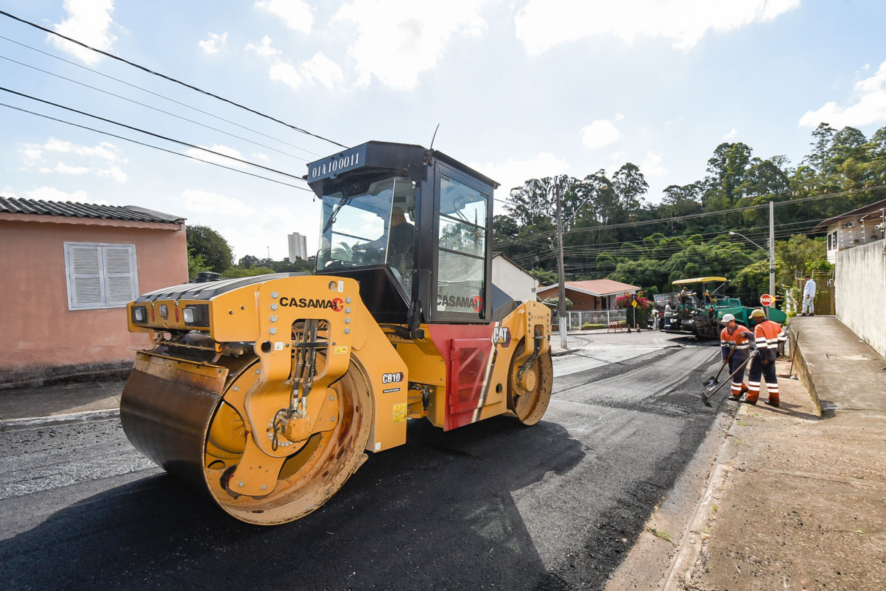 Máquina grande, amarela, passa sobre o asfalto novo na rua São Carlos Borromeu; abaixo, na mesma rua, homens espalham o asfalto; mais abaixo, outras máquinas trabalham
