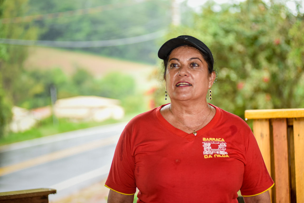 A comerciante Nilda Maria usa uma camiseta vermelha e amarela, um boné preto e está em frente da avenida Mezzalira, no Caxambú