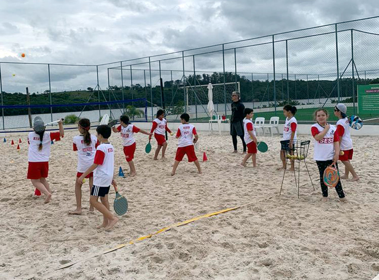 Crianças estão em quadras de areia, com raquetes em uma das mãos, aprendendo a jogar beach tennis, que é o esporte tênis em quadra de areia