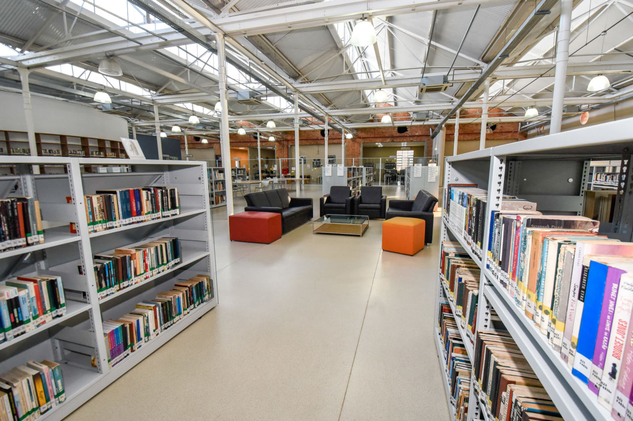 Corredor de biblioteca, com prateleiras de três andares com livros dos dois lados, com sofás e mesa ao fundo