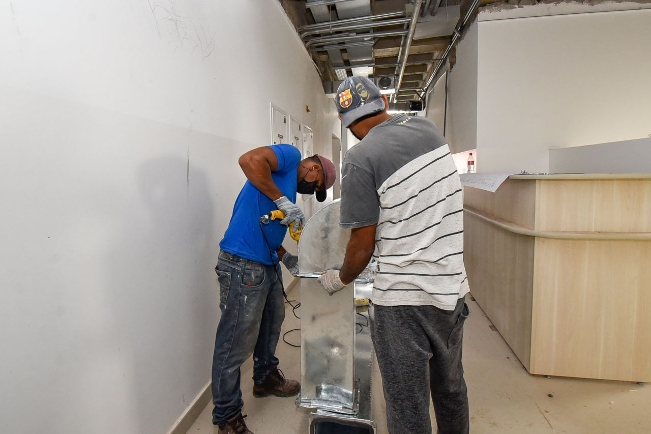 Descrição da Imagem: dois trabalhadores fazem a instalação de dutos de ventilação na Clínica da Família da Vila Hortolândia. Tubulações em metal são aparafusadas  para serem instaladas no teto do prédio, em corredor 
