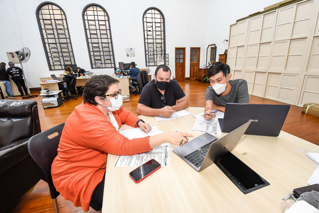 Três pessoas, com máscaras faciais, analisam e apontam para computadores e folhas sobre mesa em sala ampla com outras pessoas e armários ao fundo