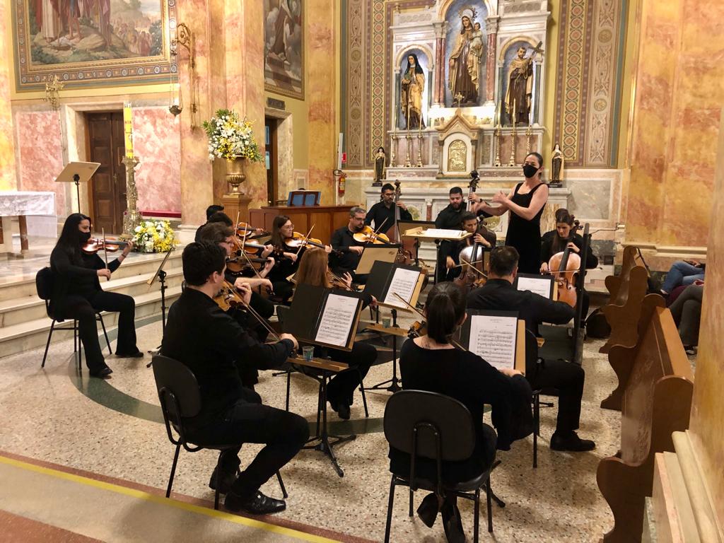 Músicos de orquestra, com roupas pretas, sentados em formato de orquestra dentro de igreja 