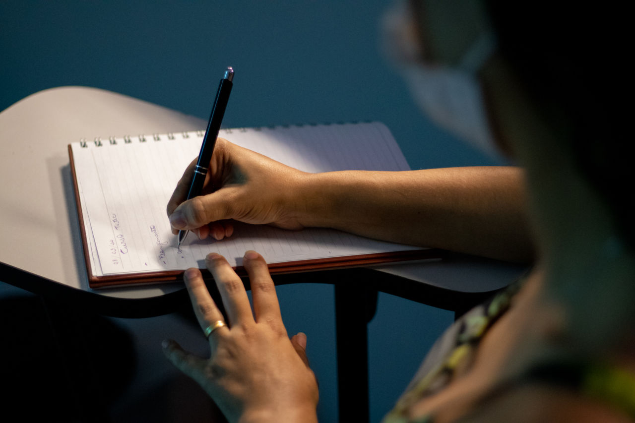 Detalhe de mulher sentada em carteira escolar, escrevendo à mão com caneta sobre caderno com pautas