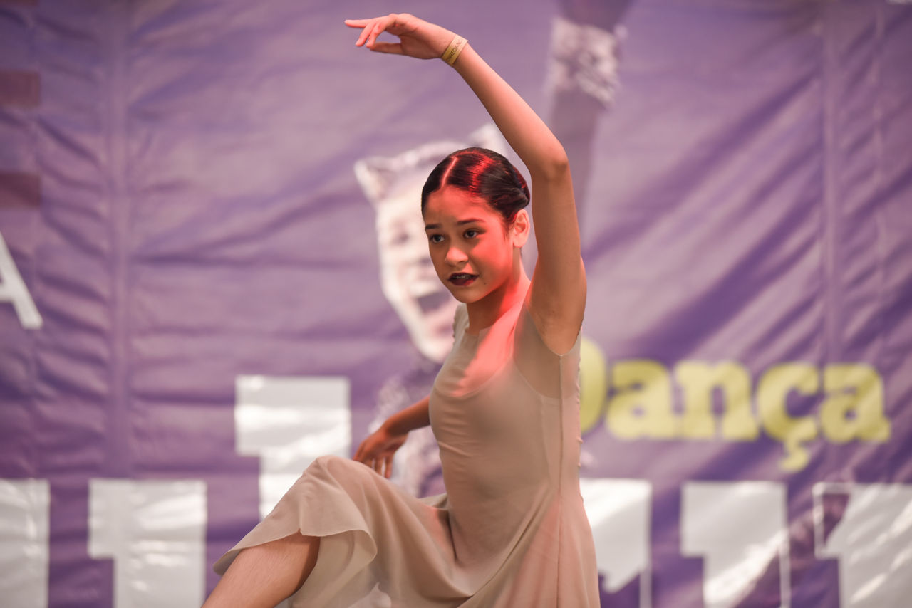 A bailarina Júlia veste bege, volta o braço esquerdo ao alto e está no palco, dançando, à frente de um banner roxo