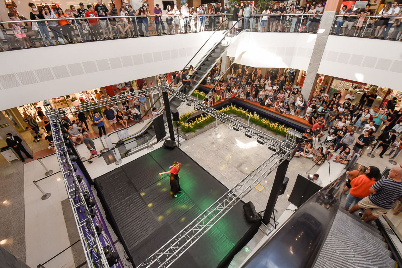 Imagem mostra o palco do Maxi Shopping de cima para baixo e pessoas assistem à apresentação de todos os lados, como no segundo piso e na escada rolante