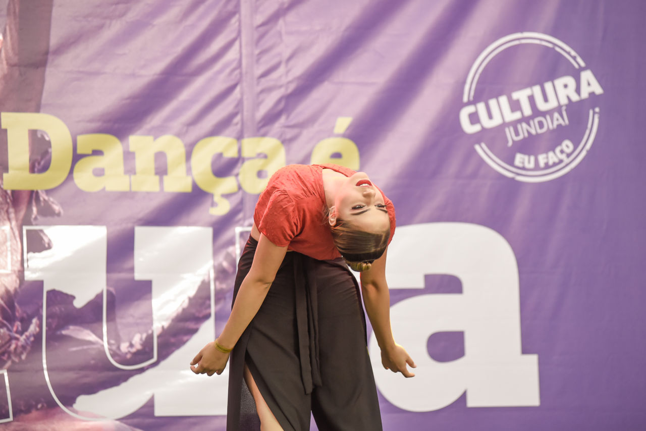 Bailarina Fernanda Martins inclina seu corpo para trás, em frente ao banner roxo do festival e sobre o palco