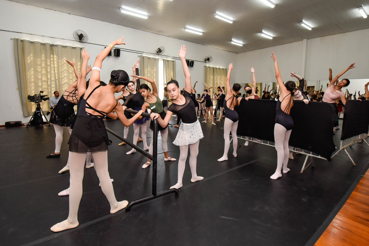 Diferentes bailarinos estão de pé, encostados na barra e dobrando seus corpos ao lado durante exercício da aula de balé