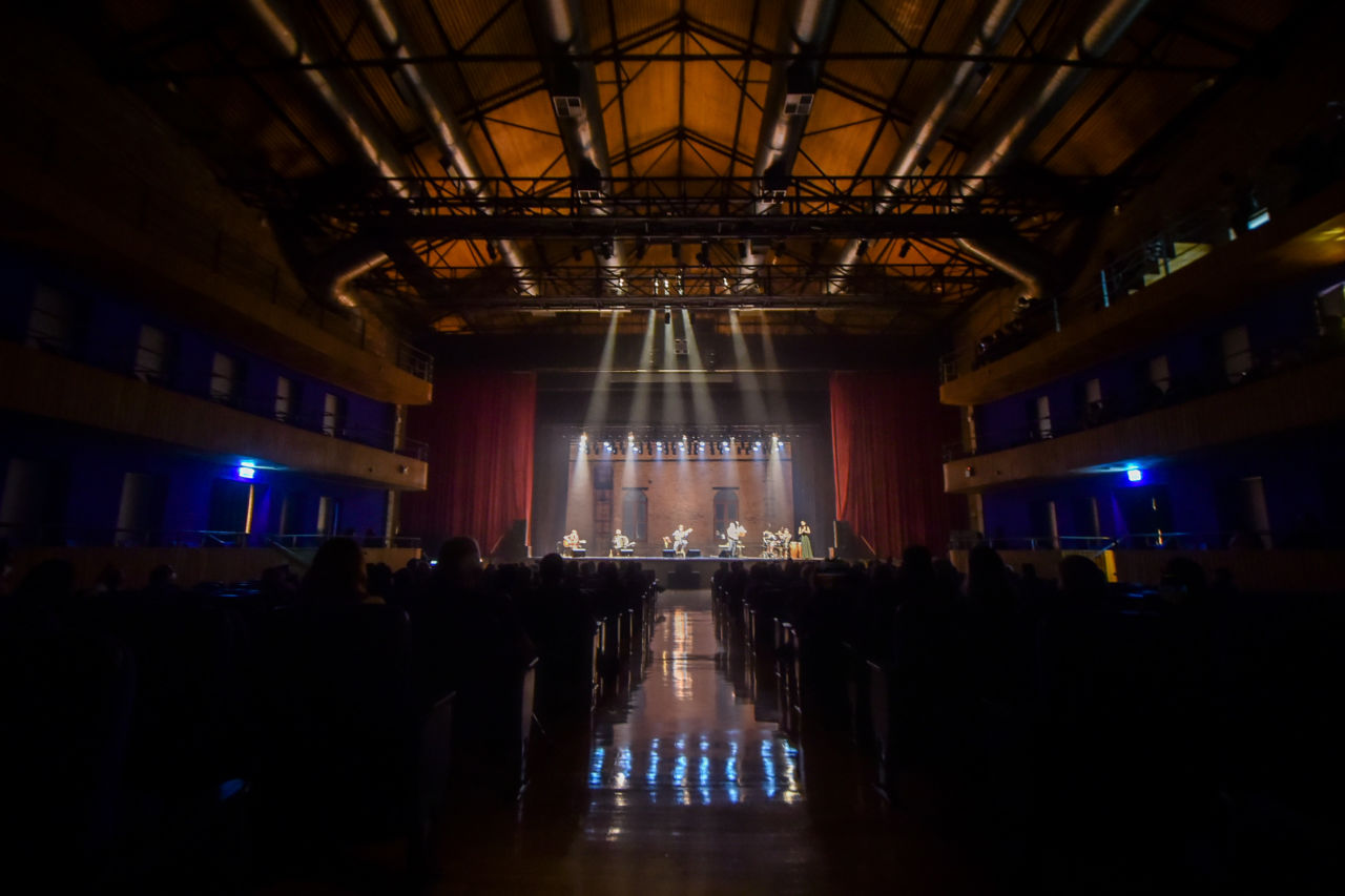 Teatro Polytheama com plateia no escuro e palco iluminado com pessoas em performance