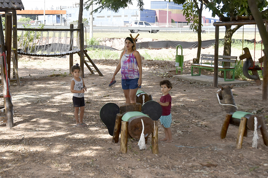 Ao centro do playground, Paula está ao lado da sobrinha e do filho, que brinca em um cavalo de madeira; ao fundo, bancos e uma ponte pequena de madeira