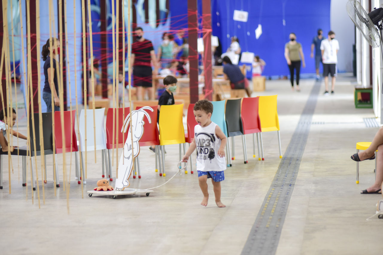 Menino de bermuda e camiseta, descalço, brinca com estrutura de madeira, puxada por uma corda, em um ambiente fechado com cadeiras coloridas, varetas de bambu e pessoas caminhando ao fundo