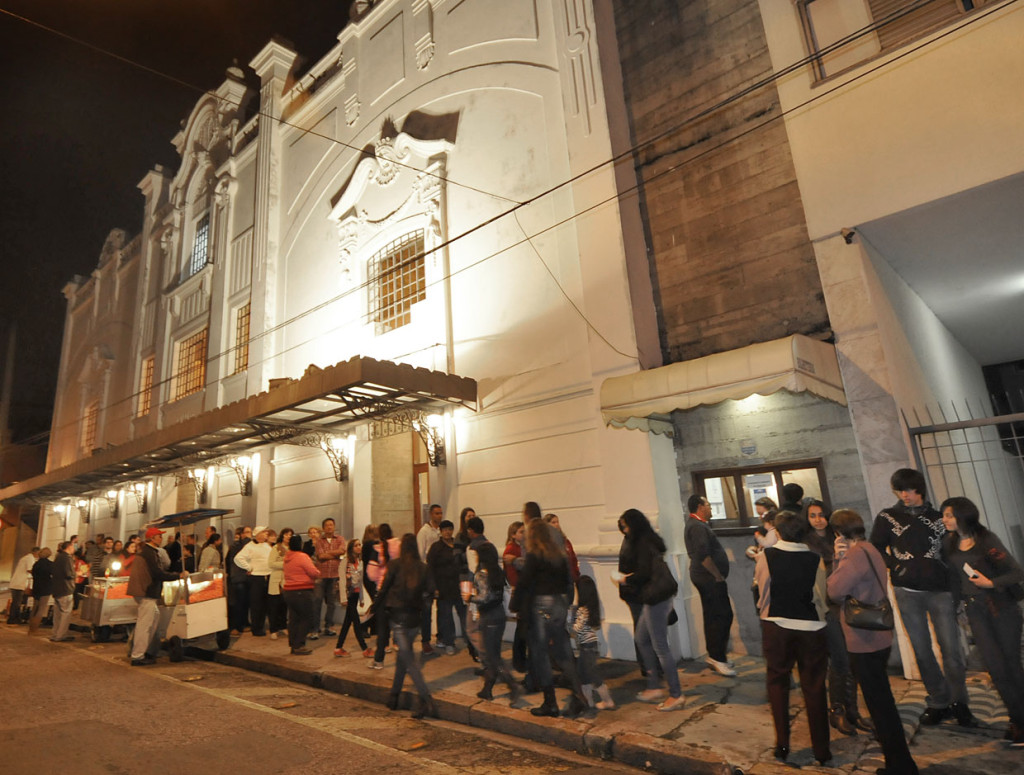 Foto noturna da fachada do teatro Polytheama iluminado, com pessoas na fila e vendedores com carrinhos de pipoca