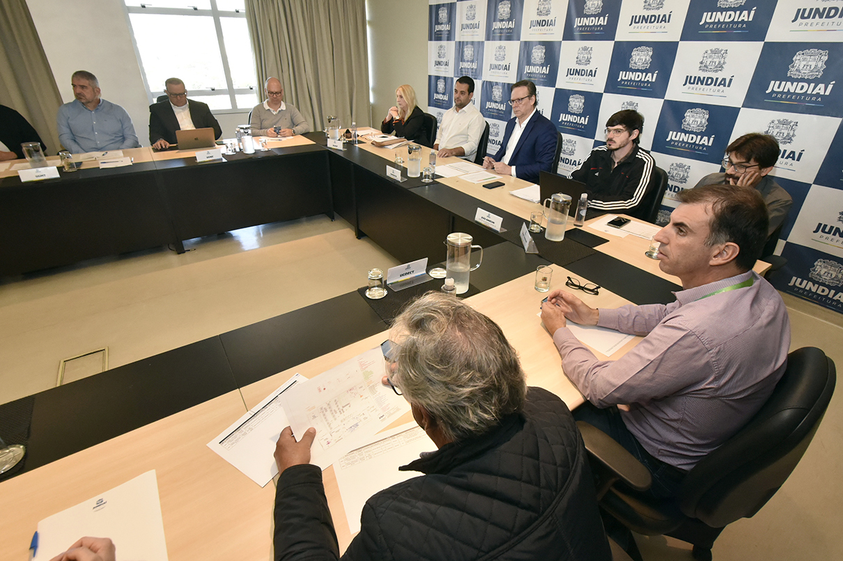 Em mesa em formato de "U", gestores conversam sobre diferentes projetos; ao fundo, um banner apresenta o logotipo da prefeitura em azul e branco