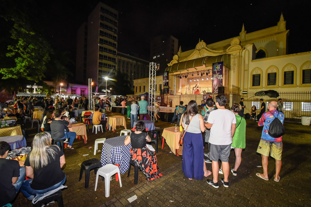Foto noturna da Praça do Coreto, com público em pé e sentado e mesas de feiras, assistindo a uma apresentação de dança sobre o palco, no fundo da igreja Matriz. Ao fundo se veem também prédios e árvores.