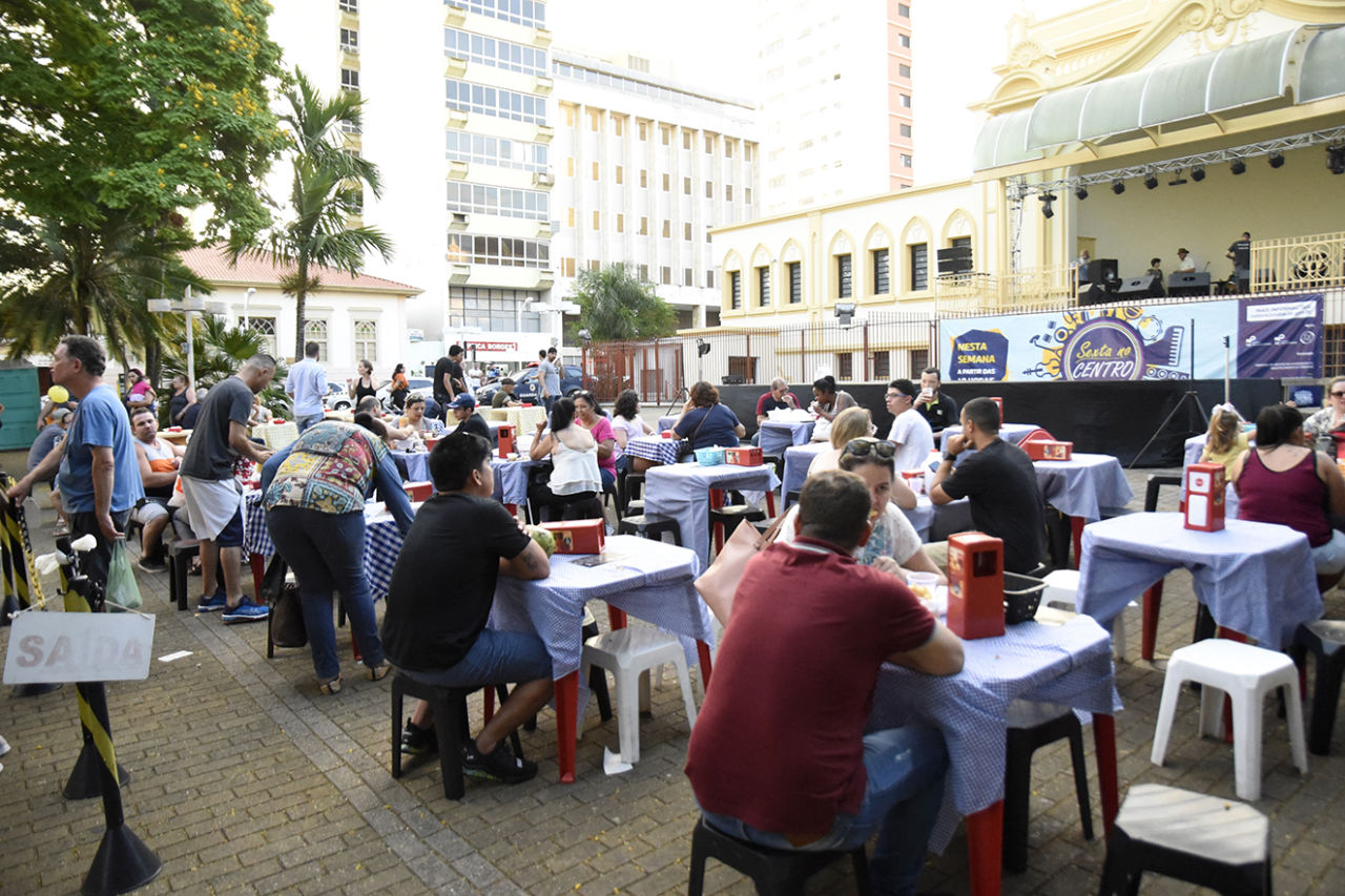 Praça do Coreto, com fundos da catedral e palco, e diversas pessoas sentadas em mesas de plástico, comendo comidas diversas