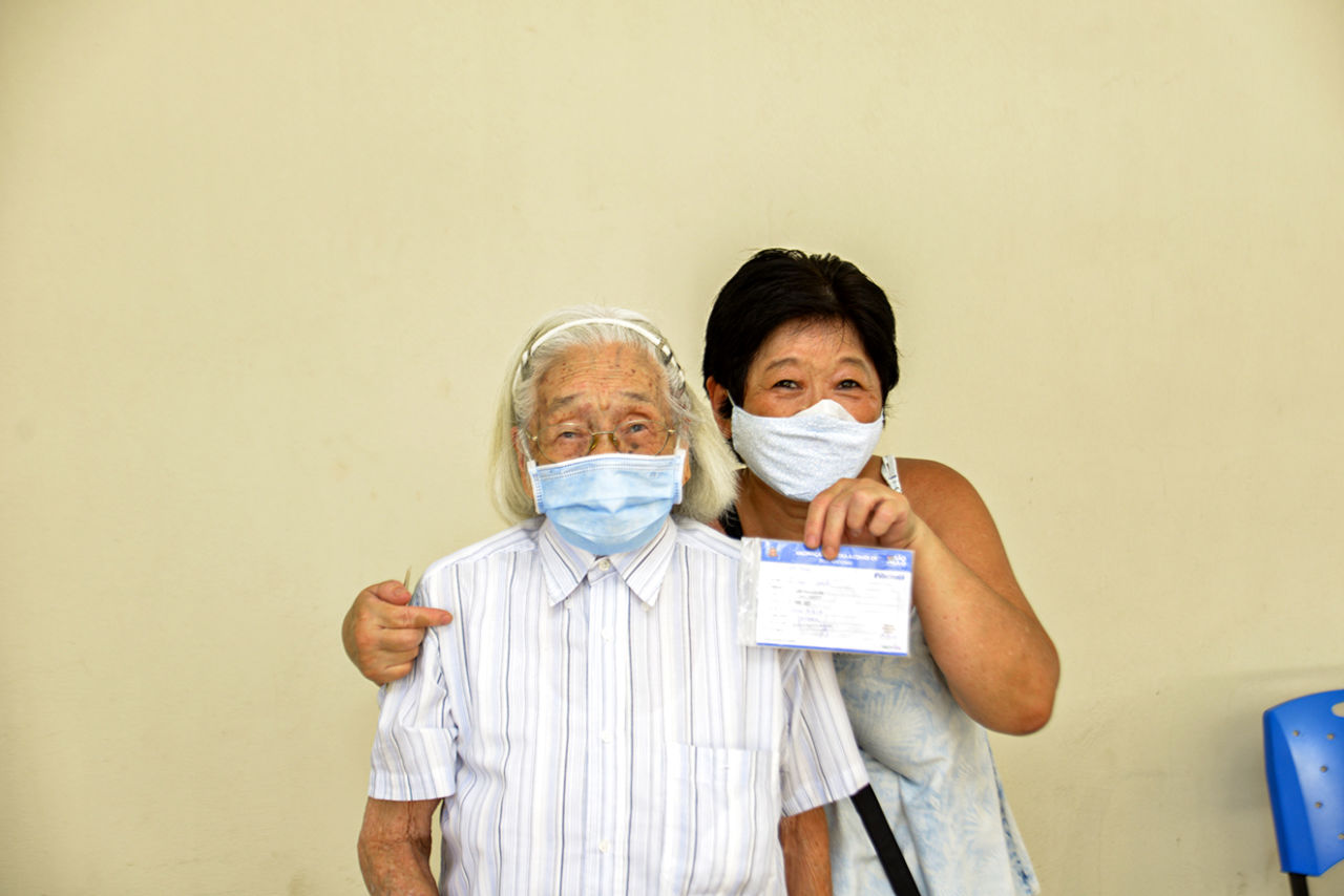 Descrição da imagem: Idosa com mais de 80 anos, de origem japonesa, usando máscara azul, camisa listada azul claro com botões, é abraçada pela filha que segura o cartão de vacinação com a mão esquerda. Ambas estão felizes com a vacinação